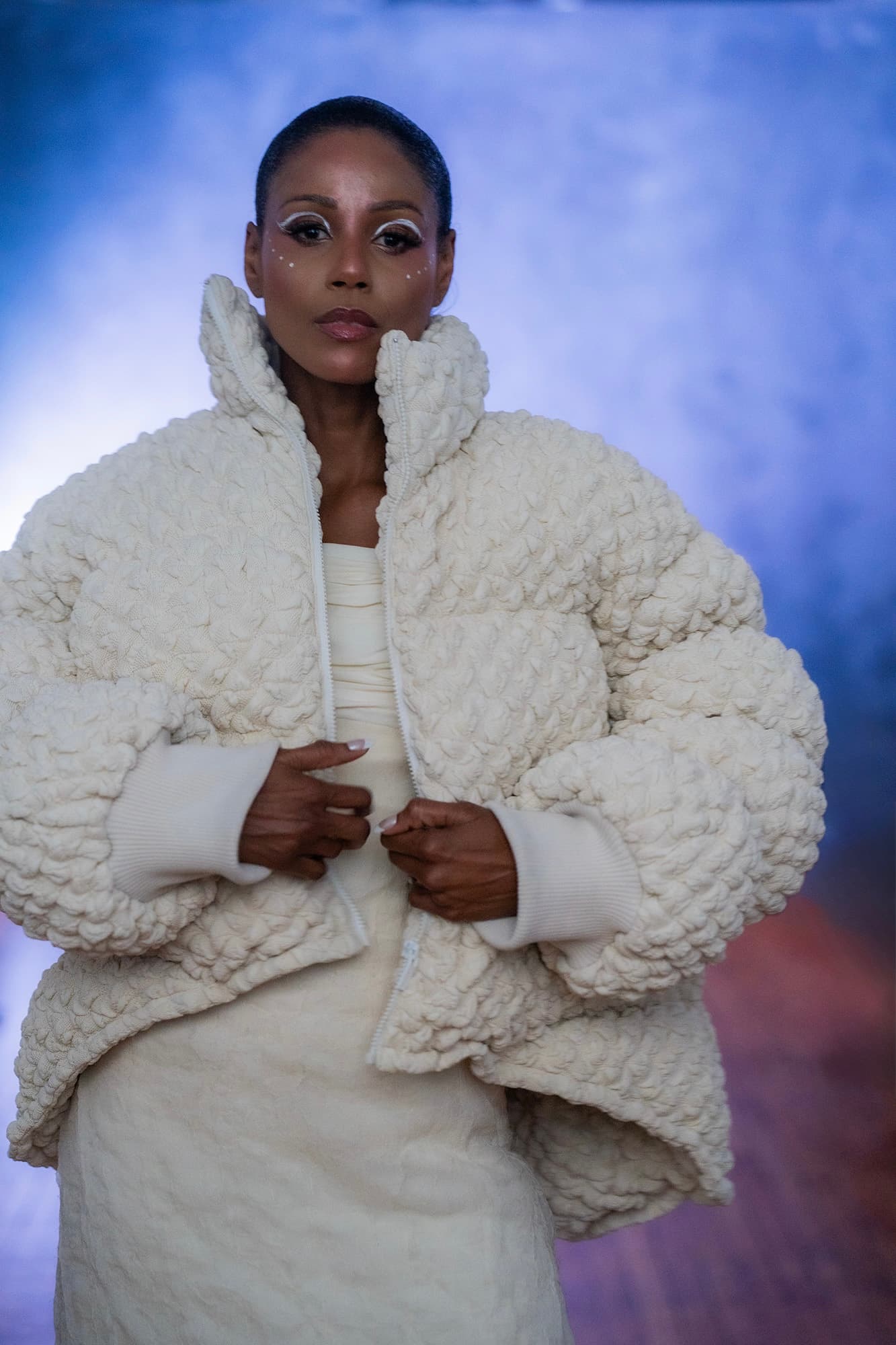 The cream bubble-knit coat close up, hands at the zip, the knit texture in full.